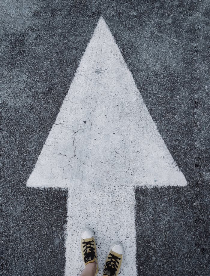 Top-down view of white arrow on asphalt with sneakers stepping forward.